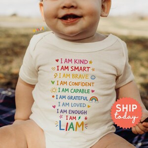 Op de afbeelding: Een cr&egrave;mekleurige babybody met kleurrijke tekst: "I AM KIND, I AM SMART, I AM BRAVE, I AM CONFIDENT, I AM CAPABLE, I AM GRATEFUL, I AM LOVED, I AM ENOUGH, I AM, LIAM". Een rode sticker zegt "SHIP today".