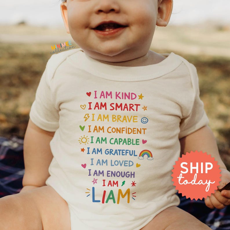 Op de afbeelding: Een cr&egrave;mekleurige babybody met kleurrijke tekst: "I AM KIND, I AM SMART, I AM BRAVE, I AM CONFIDENT, I AM CAPABLE, I AM GRATEFUL, I AM LOVED, I AM ENOUGH, I AM, LIAM". Een rode sticker zegt "SHIP today".