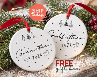 May include: Two round white ceramic ornaments with red ribbon hangers. One ornament features the black text Godmother EST. 2026 with three small pine tree icons and baby footprints. The other ornament features Godfather EST. 2026 with similar icons.