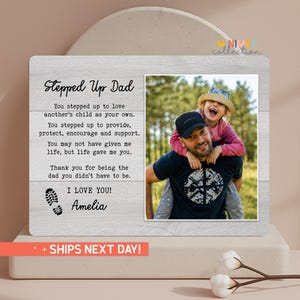 May include: A wooden photo frame with a picture of a man and a child. The frame has the text "Stepped Up Dad" and a message of appreciation. The frame is light gray with a white background.