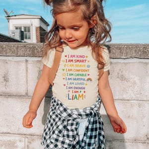 Op de afbeelding: Een jong kind draagt een cr&egrave;mekleurig t-shirt met de woorden "I AM KIND" en andere positieve affirmaties in verschillende kleuren. De naam "LIAM" staat onderaan gedrukt. Het kind draagt ook bretels en een zwart-wit geruit overhemd.
