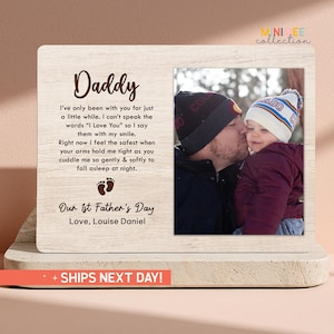 May include: A wooden photo frame with a picture of a father kissing a baby. The frame has the word "Daddy" at the top, with a poem below. The text reads "Our 1st Father's Day". The frame is on a light-colored surface.