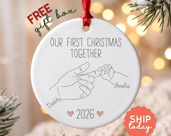 Puede incluir: Un adorno navideño redondo de cerámica blanca colgado de una cinta roja. El adorno presenta un dibujo lineal negro de una mano adulta sosteniendo el dedo de un bebé, con el texto "OUR FIRST CHRISTMAS TOGETHER", "Daddy", "Amelia" y "2026".
