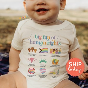 May include: A cream-colored baby bodysuit with the text "big fan of human rights" and various related graphics. The design includes phrases like "Black Lives Matter," "Love is Love," and "Feminism is for Everyone."