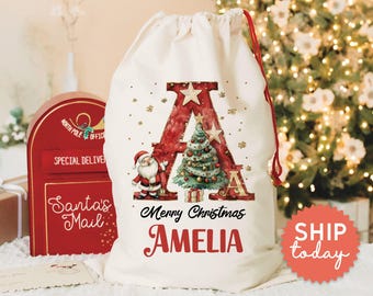 May include: A cream-colored Christmas gift bag with a red drawstring. The bag features a large red letter "A" with a Christmas tree and Santa Claus illustration. The words "Merry Christmas" and the name "AMELIA" are printed on the bag.