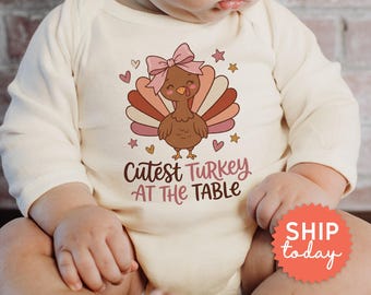 Könnte beinhalten: Ein cremefarbener Langarm-Babybody mit einer Grafik eines braunen Truthahns mit rosa Schleife, umgeben von kleinen Herzen und Sternen. Der Text "Cutest TURKEY AT THE TABLE" ist unter dem Truthahn in brauner und rosa Schrift gedruckt.
