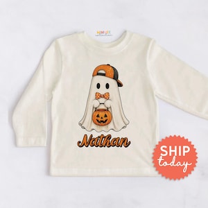 May include: A long-sleeve cream-colored shirt featuring a graphic of a ghost wearing an orange and black baseball cap and a polka-dot bow tie. The ghost holds an orange jack-o'-lantern bucket. The name Nathan is printed below in orange.
