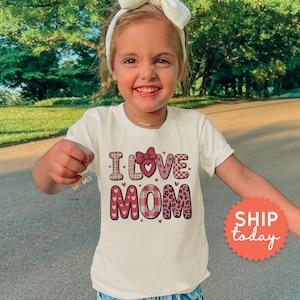 May include: A cream-colored t-shirt with the words "I Love Mom" in pink and red lettering. The shirt features a bow and heart accents. The child is wearing a white headband and smiling.