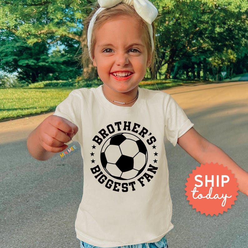 A young girl wearing a white t-shirt with a soccer ball graphic and the text "Brother's biggest fan" is smiling and pointing at the camera.
