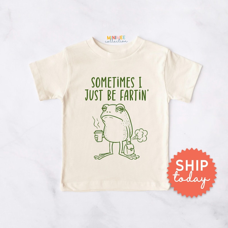 May include: Cream-colored t-shirt with a green cartoon frog graphic. The frog is holding a coffee cup and a briefcase, with the text "SOMETIMES I JUST BE FARTIN'". The shirt has short sleeves and a round neckline.