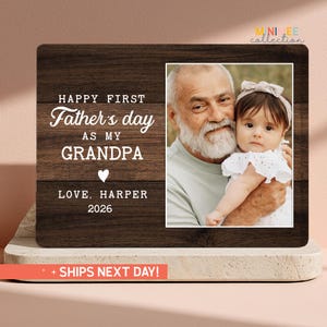 May include: A wooden photo frame with a picture of a grandfather and grandchild. The frame has the text "HAPPY FIRST Father's day AS MY GRANDPA LOVE, HARPER 2026" in white lettering. The frame is brown.