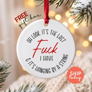 May include: A round white ceramic ornament featuring black and red text that reads: OH LOOK. IT'S THE LAST FUCK I HAVE & IT'S HANGING BY A STRING. The ornament is suspended by a thin red ribbon through a hole at the top, designed for hanging.