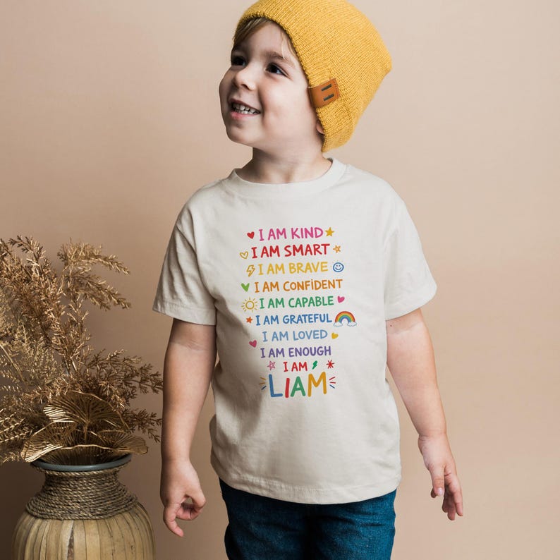 Op de afbeelding: Een beige t-shirt met kleurrijke tekst met de tekst "I AM KIND, I AM SMART, I AM BRAVE, I AM CONFIDENT, I AM CAPABLE, I AM GRATEFUL, I AM LOVED, I AM ENOUGH, I AM, LIAM". Het shirt wordt gedragen door een kind met een gele muts.