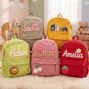 May include: Five corduroy backpacks in beige, yellow, lime green, pink, and red. Each bag features a front pocket, a top handle, and a personalized name in textured lettering. Decorative patches include a flower, a lion, cherries, and a strawberry.