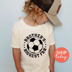 A young child wearing a white t-shirt with a soccer ball graphic and the text "Brother's biggest fan" printed on it.