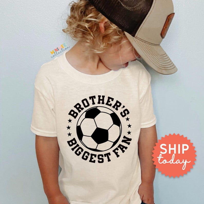 A young child wearing a white t-shirt with a soccer ball graphic and the text "Brother's biggest fan" printed on it.