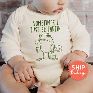 May include: Cream-colored baby onesie with long sleeves. The onesie features green text that reads "SOMETIMES I JUST BE FARTIN'" and a cartoon frog illustration holding a coffee cup and briefcase. The frog is depicted with a grumpy expression.