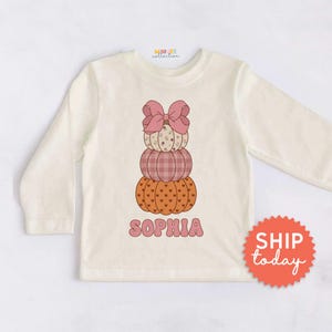 May include: A cream-colored long-sleeve shirt featuring a graphic of three stacked pumpkins topped with a large pink bow. The top pumpkin has a floral pattern, the middle has a pink plaid design, and the bottom has a heart pattern. The name SOPHIA is printed below.