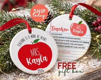 May include: Two round white ceramic ornaments with red ribbon hangers. One features a red apple graphic with the text "Teaching is a work of heart," "Mrs. Kayla," and "2026." The other shows a notebook paper design with "Teacher," "Mrs. Kayla," and definitions.