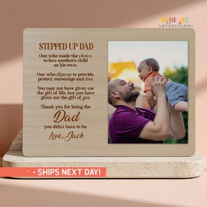 May include: A wooden picture frame with a photo of a man holding a baby. The frame has text that reads "STEPPED UP DAD" and a message of appreciation. The frame is a light brown color and has a rectangular shape.