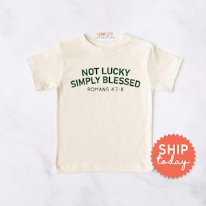 May include: Cream-colored t-shirt with the text "NOT LUCKY SIMPLY BLESSED" in green, with "ROMANS 4:7-8" below. The shirt has short sleeves and a round neckline. A red and orange sticker that says "SHIP today" is on the bottom right.