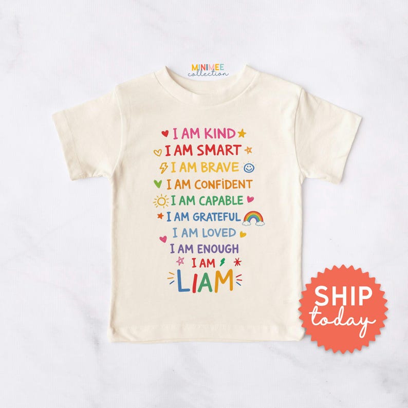 Op de afbeelding: Cr&egrave;mekleurig T-shirt met kleurrijke tekst: "I AM KIND, I AM SMART, I AM BRAVE, I AM CONFIDENT, I AM CAPABLE, I AM GRATEFUL, I AM LOVED, I AM ENOUGH, I AM, LIAM." Het shirt heeft een ronde hals en korte mouwen.