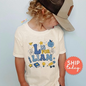 May include: A cream-colored short-sleeve t-shirt featuring a blue and yellow graphic print that reads L IS FOR LIAM. The design includes illustrations of an apple, a pencil, a book, a lightbulb, a crayon, stars, and hands making peace signs.