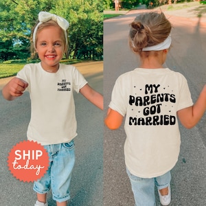 May include: Off-white short-sleeved t-shirt with the text "My Parents Got Married" on the front and back. The shirt is worn by a young child wearing blue jeans and a white bow. The shirt is a casual style.