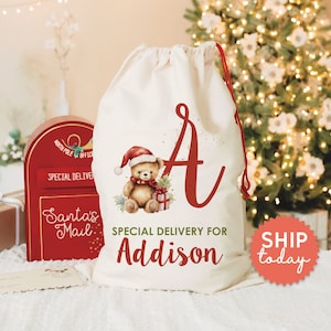 May include: A white canvas Santa sack with a red drawstring. The sack has a red letter "A" and a watercolor illustration of a teddy bear wearing a Santa hat and holding a gift. The text "SPECIAL DELIVERY FOR Addison" is printed below the illustration.