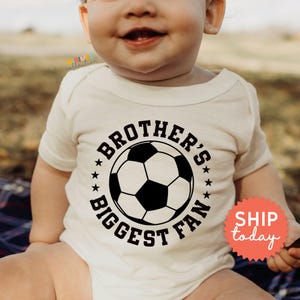 A smiling baby wearing a white t-shirt with a soccer ball graphic and the text "BROTHER'S BIGGEST FAN" printed on it.