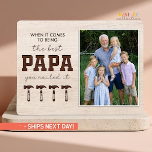May include: A wooden photo frame with a family portrait and the words "WHEN IT COMES TO BEING the best PAPA you nailed it." The names Amelia, Harper, Liam, and Henry are below the text. The frame is on a light-colored surface.