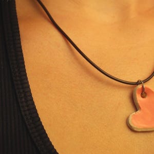May include: A pink heart pendant necklace with a black cord. The heart-shaped pendant has a light pink center and a white border. The necklace is worn against a black top.