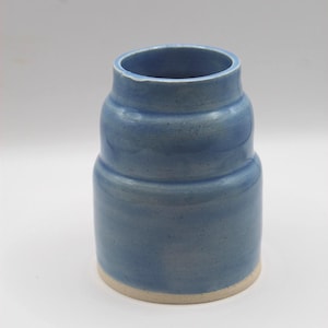 May include: A handmade, blue ceramic vase with a cylindrical shape and a slightly flared top. The vase has a light blue glaze with subtle variations in color and texture. The base is a natural, unglazed beige.