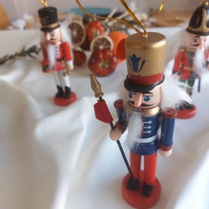 Könnte beinhalten: Holz-Nussknacker-Ornamente in Rot, Blau und Gold. Die Ornamente haben die Form von Soldaten mit detaillierten Uniformen und Hüten. Jedes Ornament hat eine goldene Schnur zum Aufhängen. Der Hintergrund umfasst getrocknete Orangenscheiben und ein weißes Tuch.