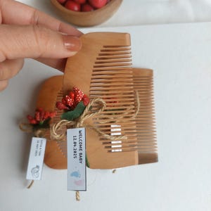 May include: Two wooden combs tied with twine and adorned with red berry sprigs. Each comb has a tag that reads "WELCOME BABY 12.04.2025" and "SOFIA & TOM 28 MAY 2025". The combs are light brown in color.
