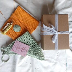 Pode incluir: Uma bolsa de crochê laranja com um pequeno círculo verde e um chaveiro que diz "Louise". Um triângulo de crochê verde e um cartão de aniversário rosa também estão presentes. Uma caixa de presente marrom é amarrada com uma fita branca.