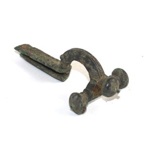 May include: An antique bronze brooch, featuring a curved, arched design with decorative detailing. The brooch has a pin and a catch, and two round decorative elements at the end. The metal has a weathered, aged appearance.