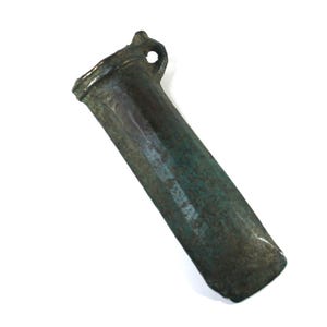May include: A weathered bronze-coloured metal socketed axe head featuring a cylindrical socket with a small circular loop on one side. The surface displays a dark, oxidised patina with visible green and brown tones throughout the elongated body.