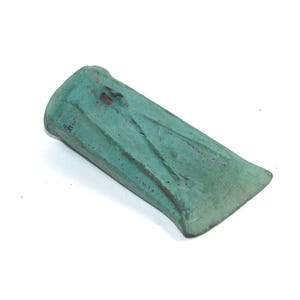 May include: A weathered, teal-coloured metal wedge tool, possibly for splitting wood. The wedge has a rectangular shape with a beveled edge and a slightly curved top. The surface shows signs of age and use, with a textured appearance.