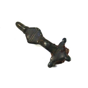 May include: A dark, weathered metal brooch with a T-shaped design, featuring a central diamond-shaped section, a slender stem, and a crossbar with three rounded protrusions at the base, showing signs of oxidation and age-related surface wear.