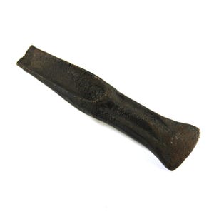 May include: A dark brown, weathered metal chisel with a textured, pitted surface. The tool features a tapered, elongated body with a wide, flat striking end and a narrow, sharpened blade edge at the opposite side, showing signs of age and heavy use.