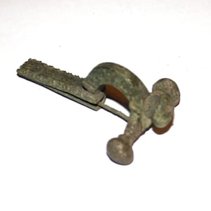 May include: An antique bronze brooch with a curved pin and a decorative head. The brooch has a rectangular clasp with a textured edge. The metal has a weathered, aged appearance, suggesting it is an old piece.