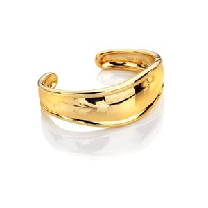 May include: A gold-colored cuff bracelet with a smooth, curved design. The bracelet has a small, embedded gemstone and reflects light, creating a shiny appearance. The jewelry is set against a white background.