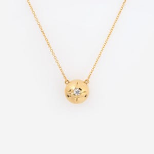 May include: A gold-colored necklace with a round pendant. The pendant features a starburst design with a small, clear gemstone at its center. The chain is delicate and also gold-colored.