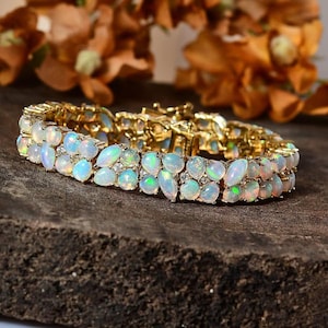 Può includere: Un bracciale d'oro con multipli opali. Gli opali sono ovali e rotondi, con una miscela di colori bianco, blu e verde. Il bracciale è su una superficie marrone scuro, con fiori arancioni sfocati sullo sfondo.