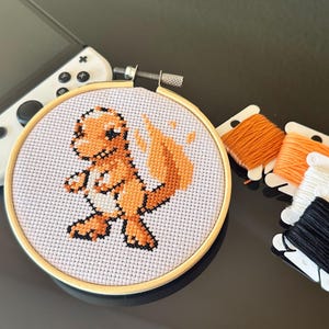 Könnte beinhalten: Ein Kreuzstich-Stickrahmen mit weißem Hintergrund und einer orange-braunen Comicfigur mit Flammenschwanz. Der Rahmen ist von orangem und schwarzem Stickgarn umgeben.