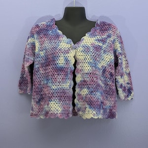 May include: A crocheted cardigan with a tie-dye pattern in shades of purple, blue, and white. The cardigan has a scalloped hem and a V-neck.