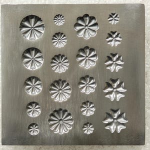 May include: A square metal plate featuring a grid of embossed floral shapes in varying sizes. The design includes multiple circular flower patterns with radiating petals and several four-petaled flower motifs arranged in columns and rows.