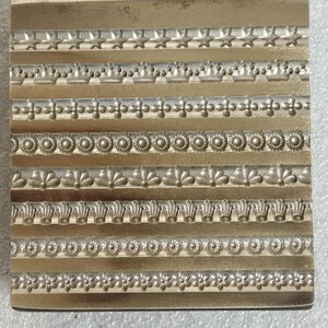 Puede incluir: Un sello metálico dorado rectangular con múltiples diseños de bordes decorativos. Los bordes incluyen estrellas, coronas, patrones florales y otros elementos ornamentados. El sello se utiliza probablemente para crear diseños en relieve en papel u otros materiales. Mide aproximadamente 10 cm x 10 cm.