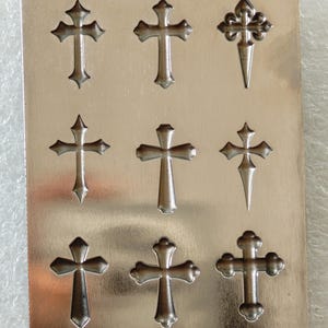 May include: A metal sheet featuring nine embossed crosses in different styles. The crosses are silver-coloured and arranged in three rows of three. The designs include classic and decorative cross shapes.